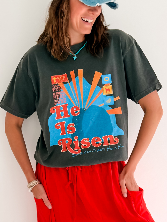 He Is Risen tee in dark gray, featuring colorful graphic design and a relaxed fit, worn with red shorts.