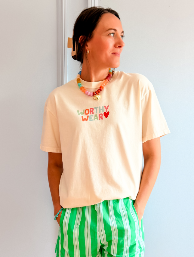 Cream-colored tee with "Worthy Wear" text, worn by a woman with green striped shorts and colorful necklace.