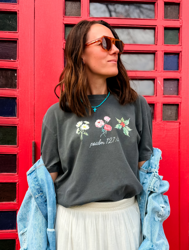 Custom birth flower tee in dark gray with floral designs, worn by a woman with sunglasses against a red background.