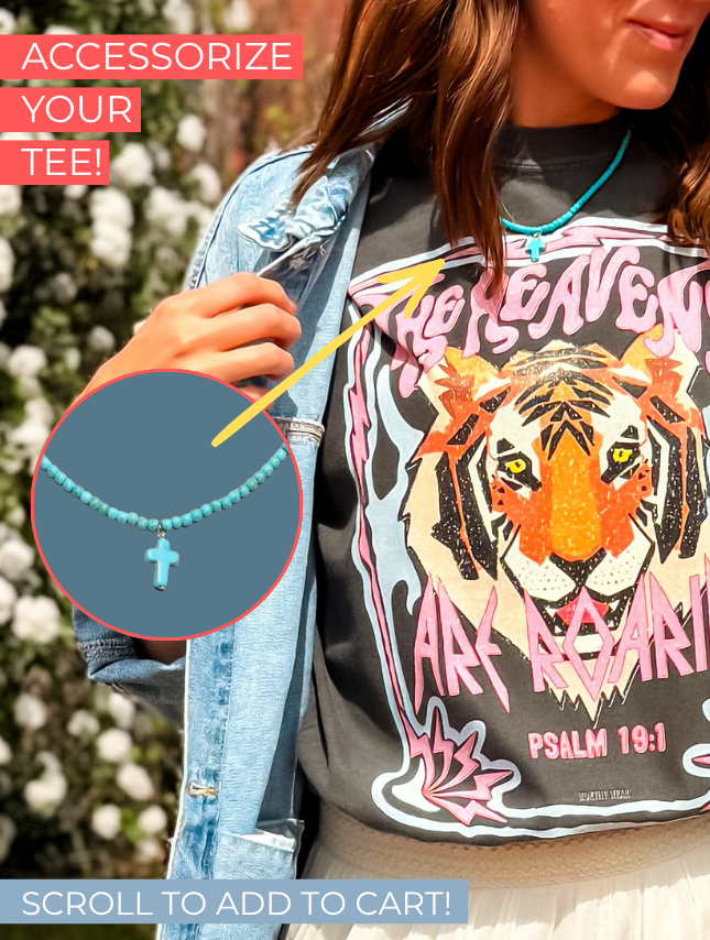 The Heavens Are Roaring tee features a graphic of a tiger with colorful text, worn by a person with a turquoise necklace.