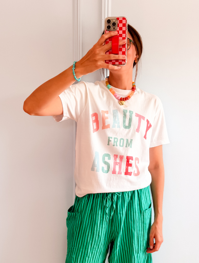 Beauty From Ashes tee in white with colorful text, worn by a person holding a phone, paired with green striped pants.