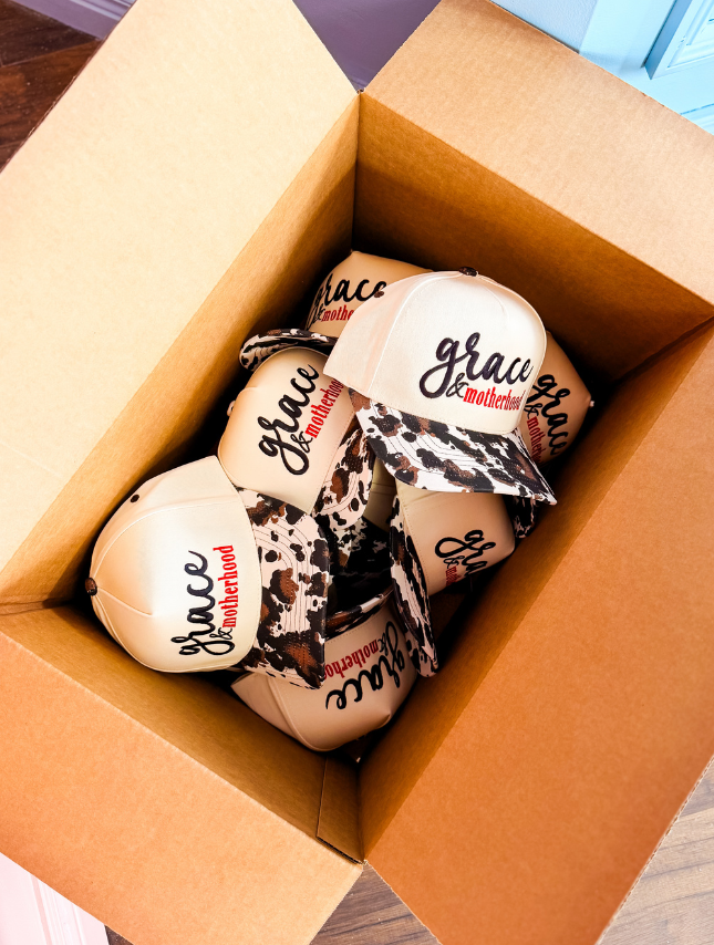 Grace & Motherhood trucker hats in a cardboard box, featuring beige caps with black and red text and leopard print brims.