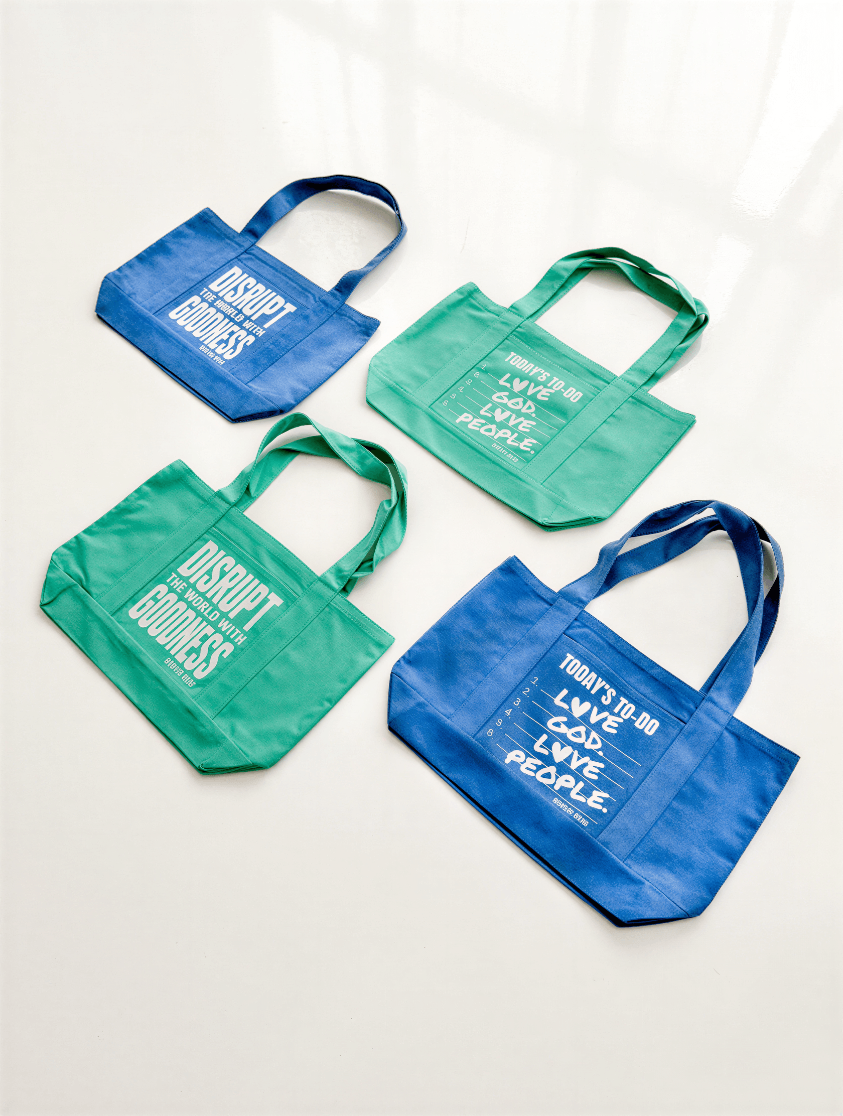 Disrupt The World tote in sea green with text designs, displayed alongside three other tote bags in varying shades of green.