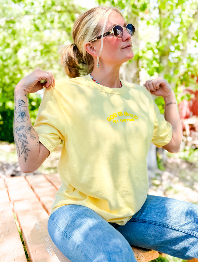 Yellow God Is Good Puff Tee worn by a woman sitting outdoors, with sunglasses and a tattoo visible on her arm.
