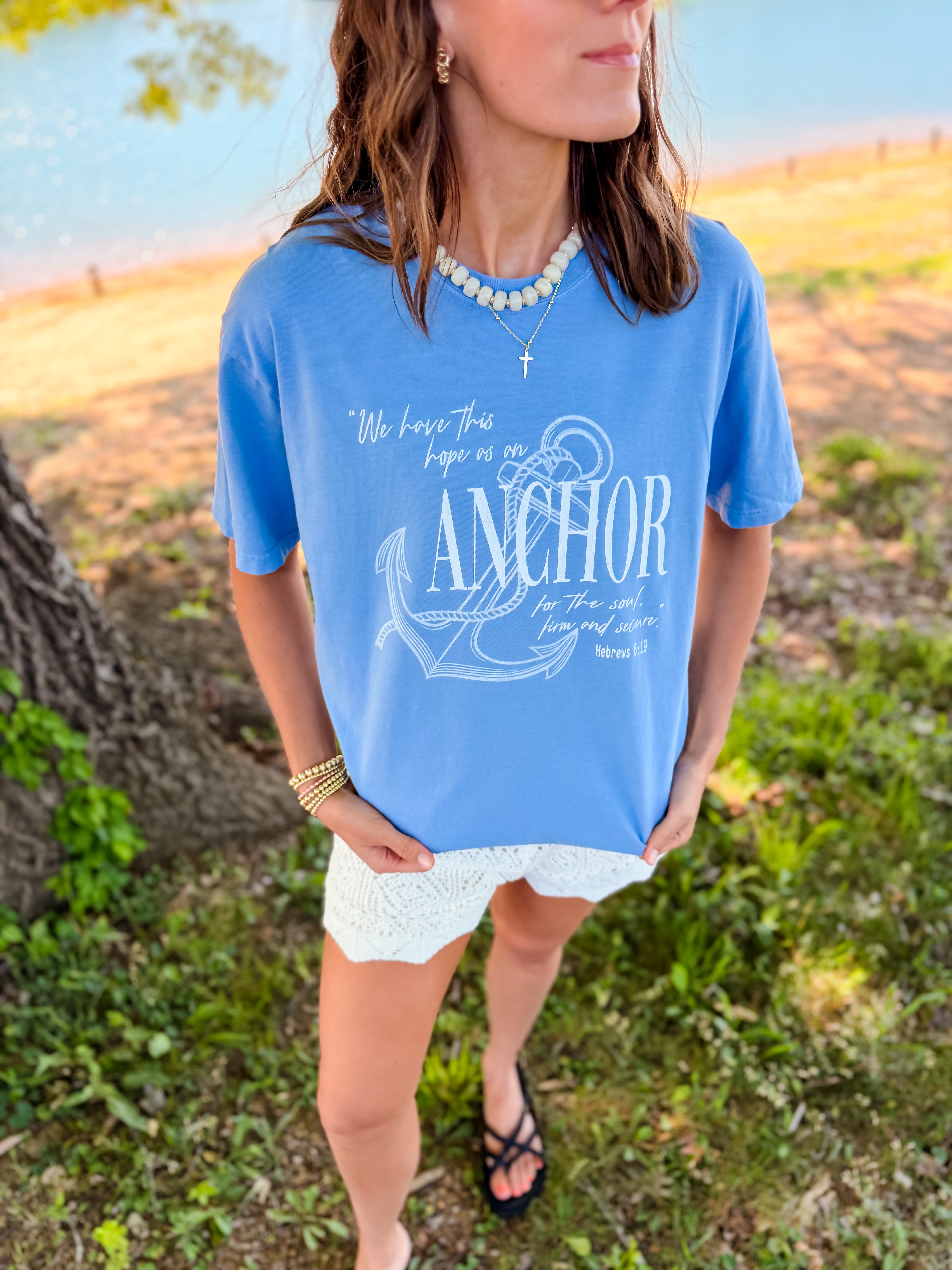Blue Anchor for the Soul tee with white graphic text, worn by a model near a tree by a lake, paired with white shorts.