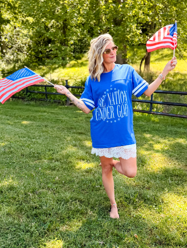 Blue Under God jersey tee with white lace trim, worn by a woman holding American flags in a grassy outdoor setting.