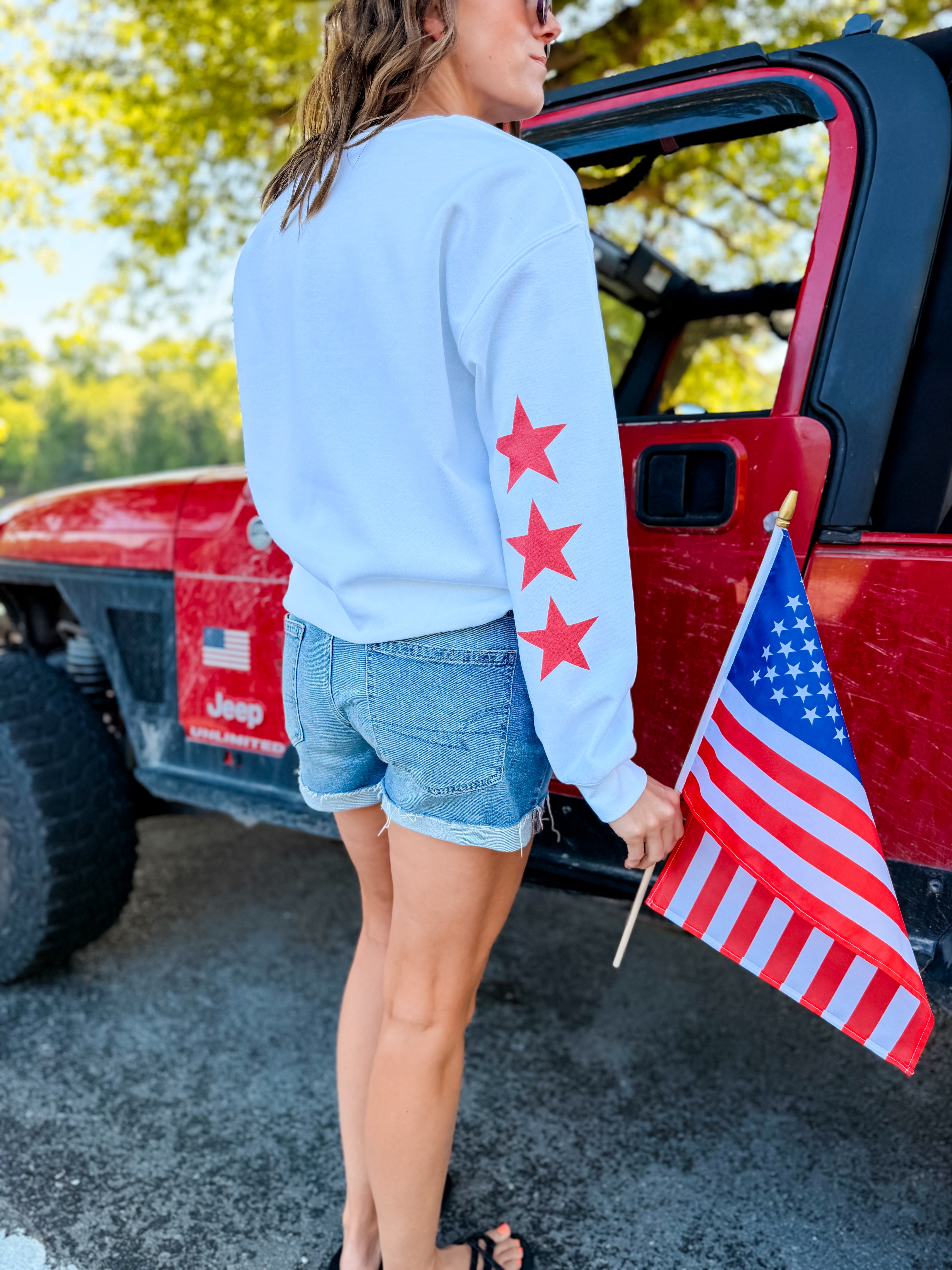 White sweatshirt featuring red stars on the sleeve, paired with denim shorts and holding an American flag.