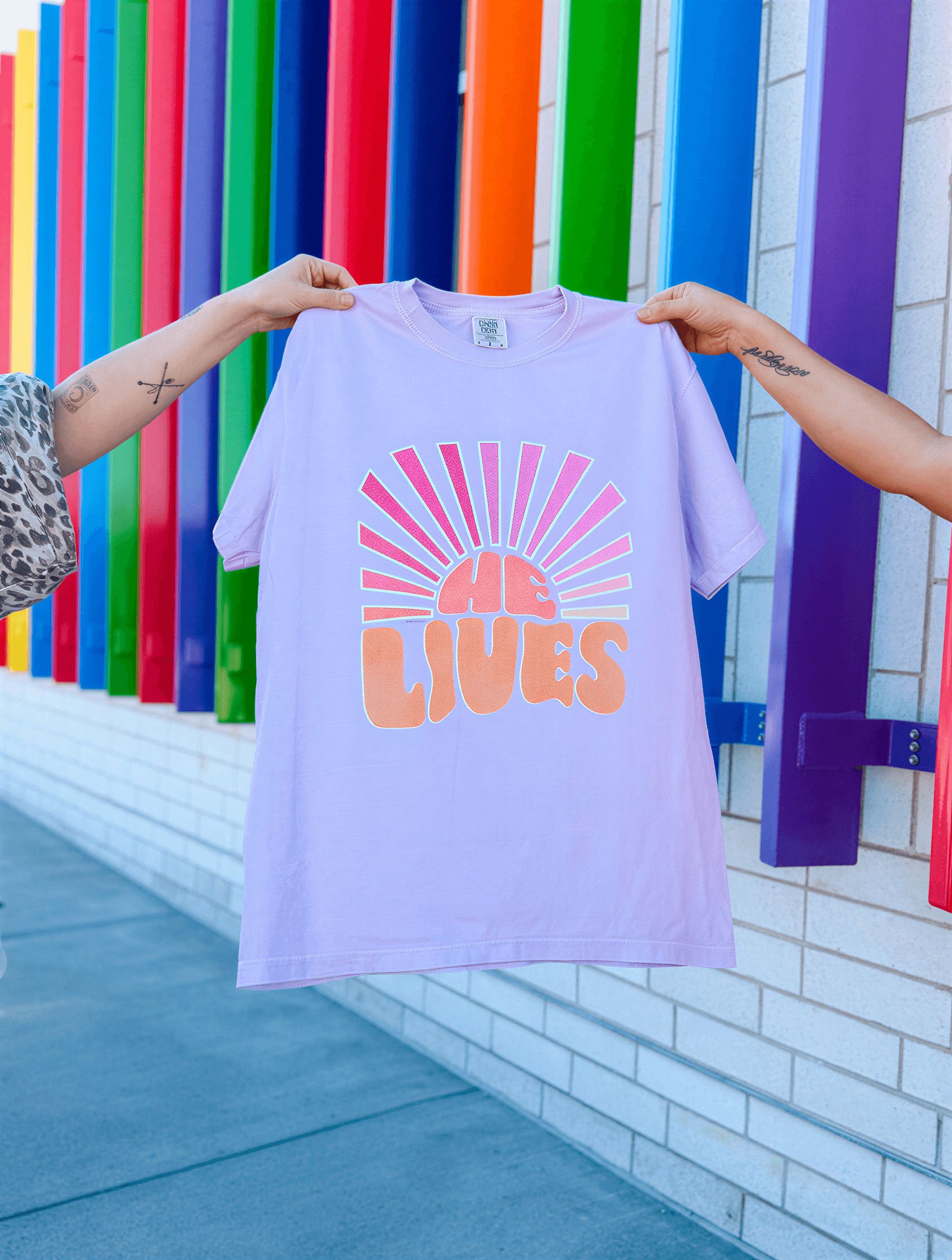 Person holding a purple t-shirt with 'HE LIVES' design against a colorful background