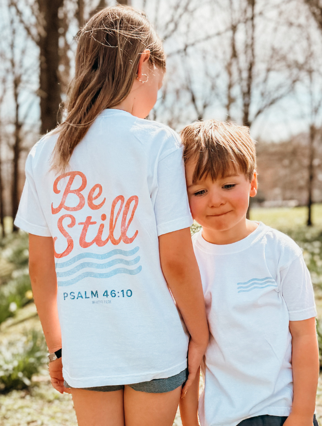 Youth tee featuring "Be Still" text and Psalm 46:10, worn by a girl and boy outdoors in a park setting.