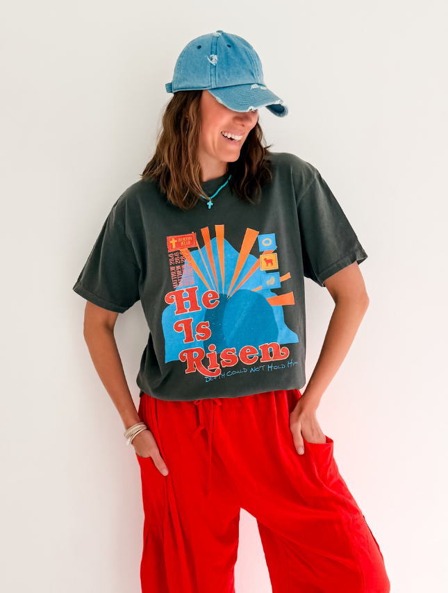 He Is Risen tee in black with colorful graphic design, paired with red pants and a blue cap, worn by a smiling model.