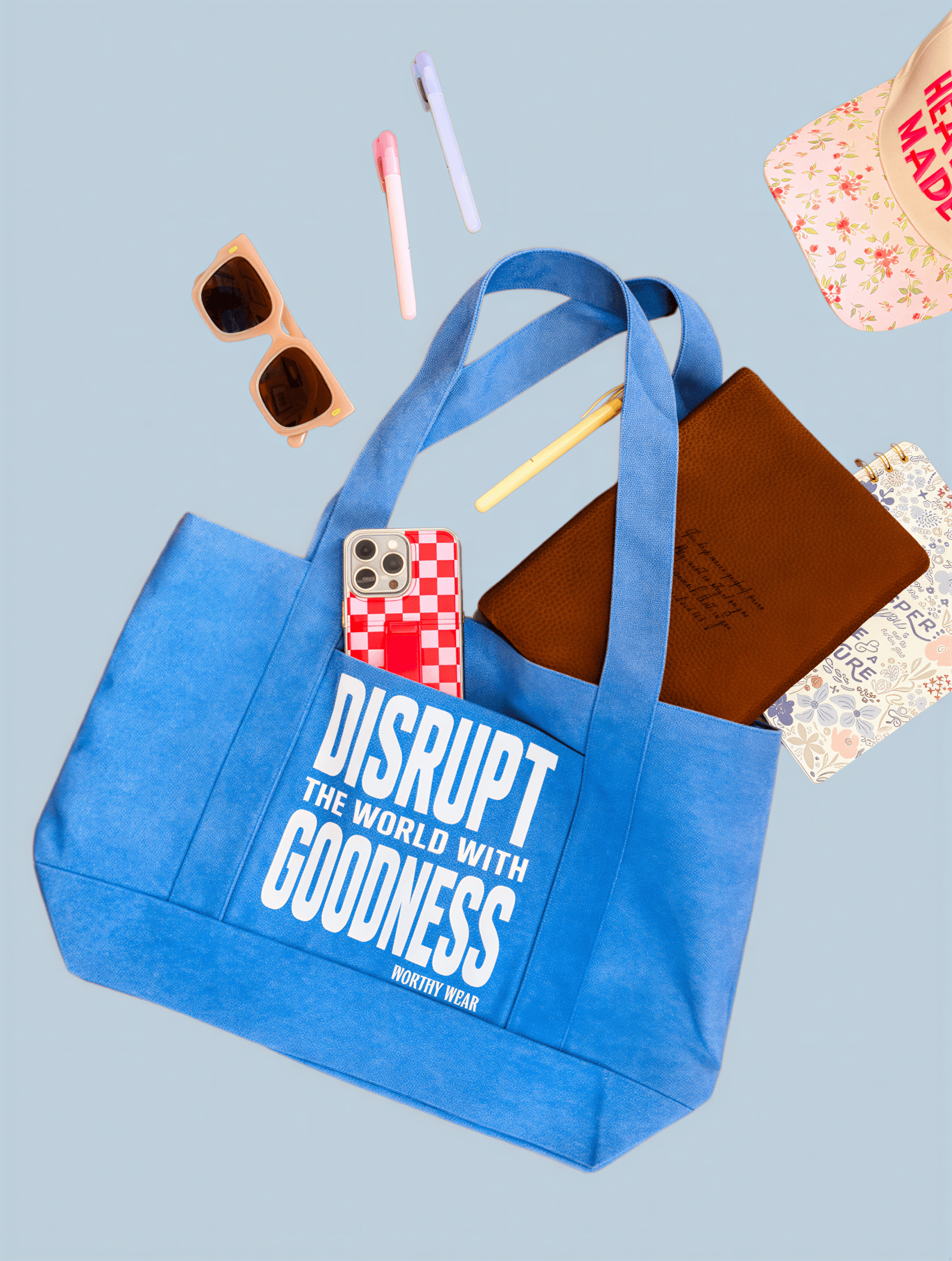 Disrupt the World tote in periwinkle blue with white text, surrounded by sunglasses, a notebook, and pens.