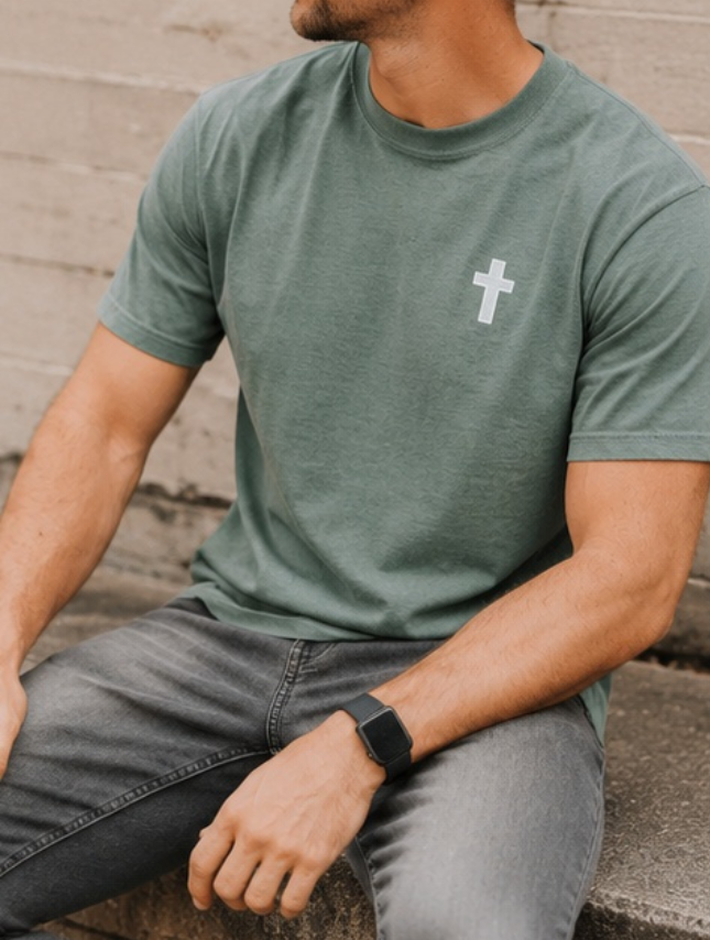 He Moves Mountains tee in moss green with a white cross design, worn by a man sitting on a stone surface.