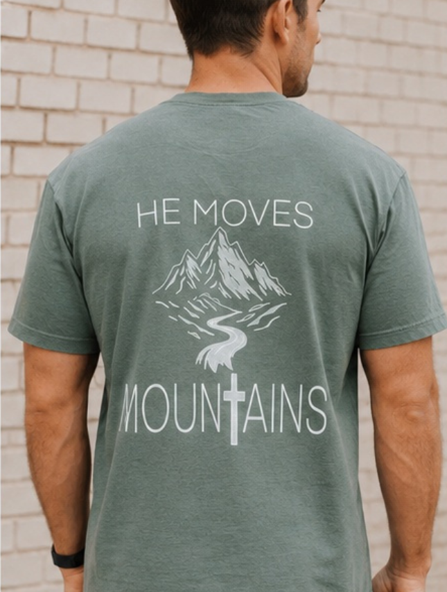 He Moves Mountains tee in moss green, featuring a mountain graphic and cross design on the back.