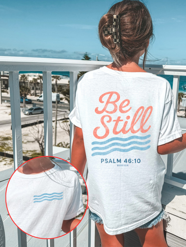 Person wearing a white t-shirt with 'Be Still' text on a balcony with ocean view