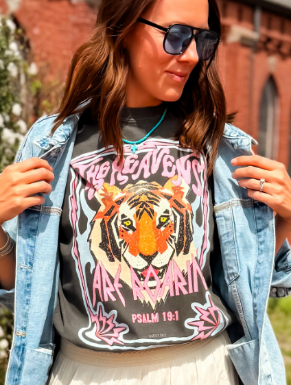 The Heavens Are Roaring tee features a tiger graphic with pink and black accents, worn by a woman in a denim jacket.