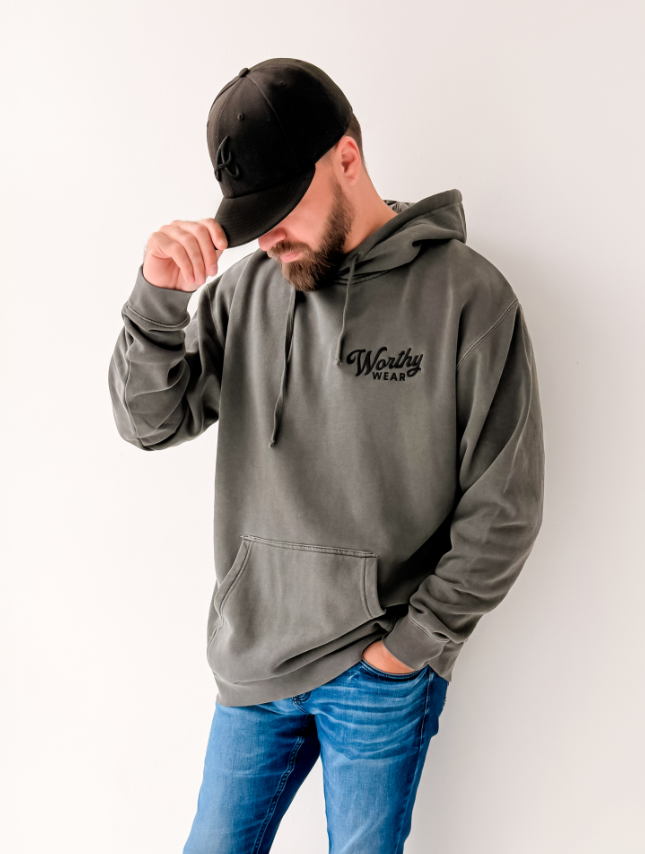 Worthy Wear washed hoodie in dark gray, featuring a front pocket and embroidered logo, worn by a man with a black cap.