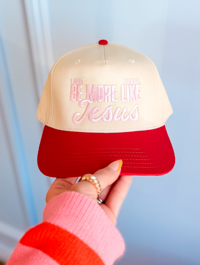 More Like Jesus trucker hat with cream front, red brim, and pink embroidered text. Handheld against a light background.