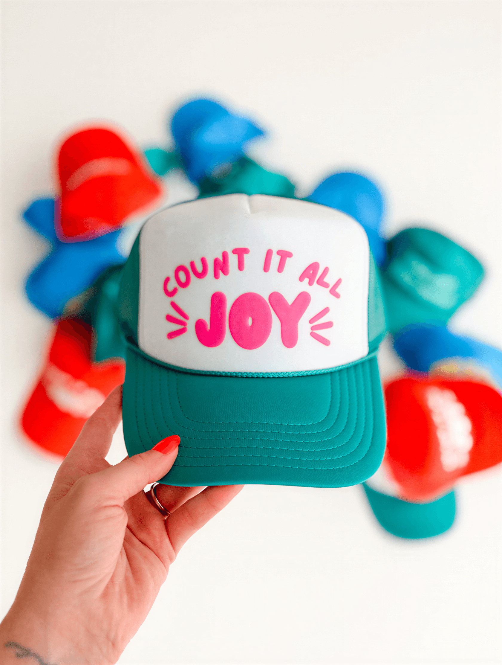 Count It All Joy foam trucker hat in jade with pink text, held in hand, surrounded by colorful caps.