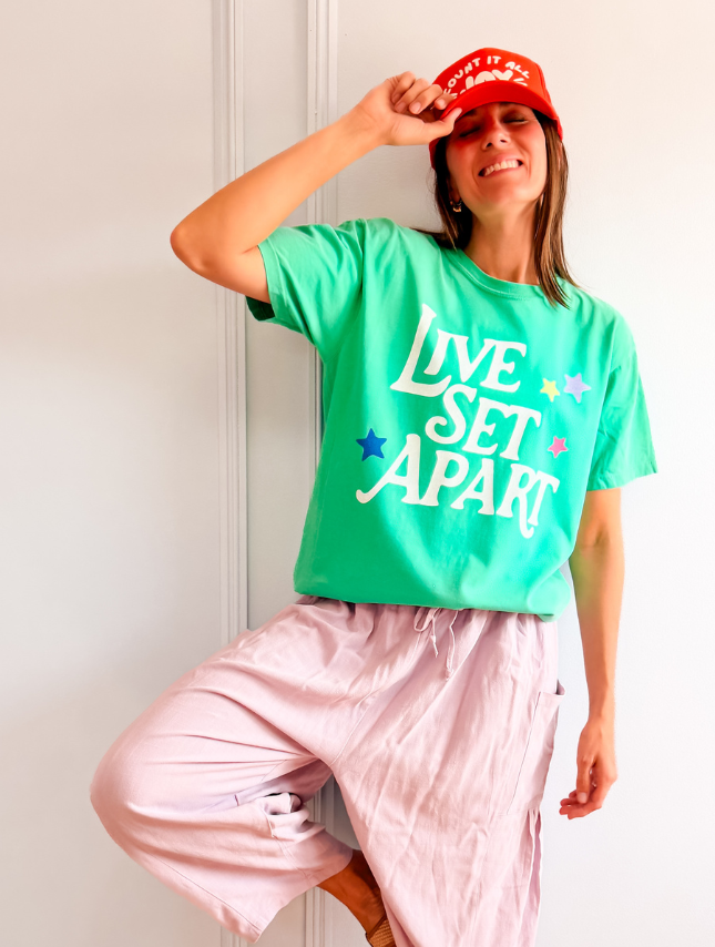 Green Live Set Apart tee with white text and colorful stars, worn with light pink pants and a red cap.