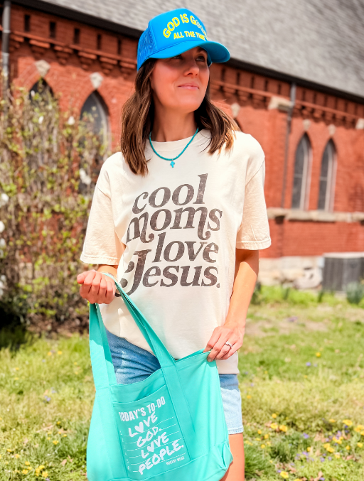 Cool Moms Love Jesus tee in cream color, worn by a woman with a blue cap, holding a turquoise tote bag outdoors.