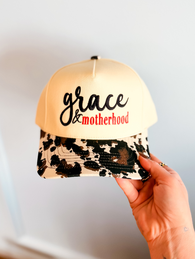 Grace & Motherhood trucker hat with a cream front and black and brown patterned brim, held in a hand.
