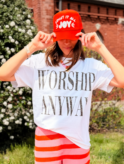 Person wearing a white t-shirt with 'Worship Anyway' text, red cap, and red and white striped shorts outdoors.