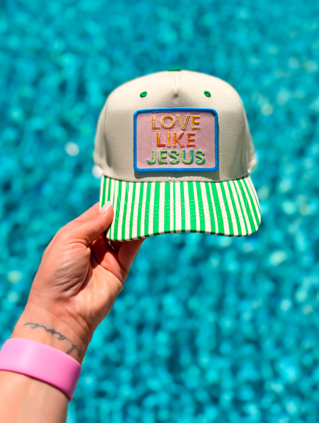 Love Like Jesus trucker hat with a beige front and green striped brim, held against a blue water background.