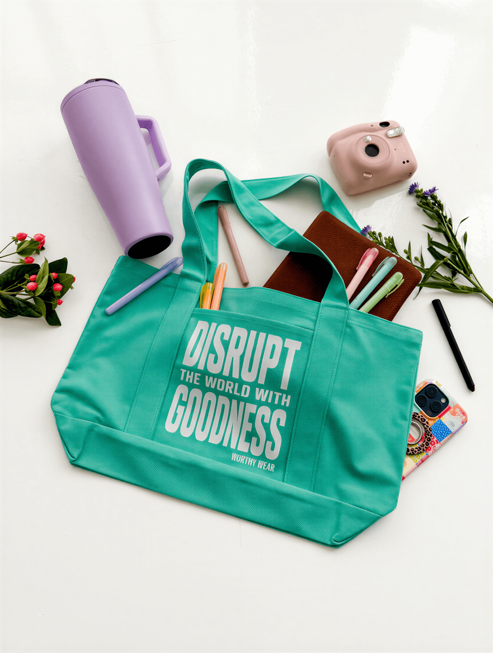 Sea green tote bag with "Disrupt the world with goodness" text, surrounded by pens, a notebook, and a camera.
