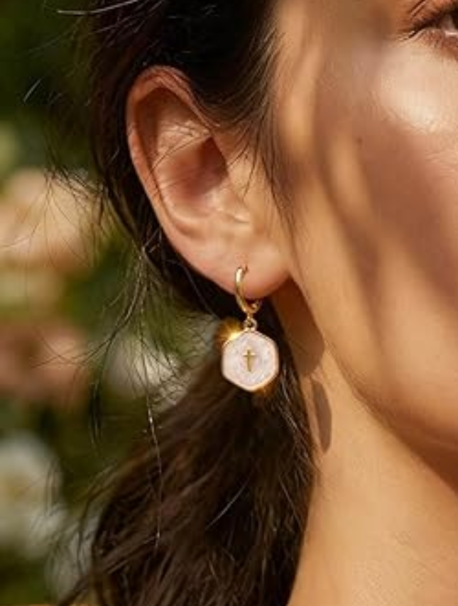 White pearl cross drop earrings with gold accents, worn on a model's ear against a blurred floral background.