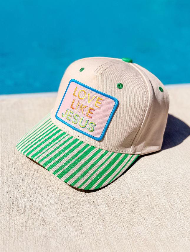 Love Like Jesus trucker hat in beige with a colorful patch and green striped brim, placed on a light surface.