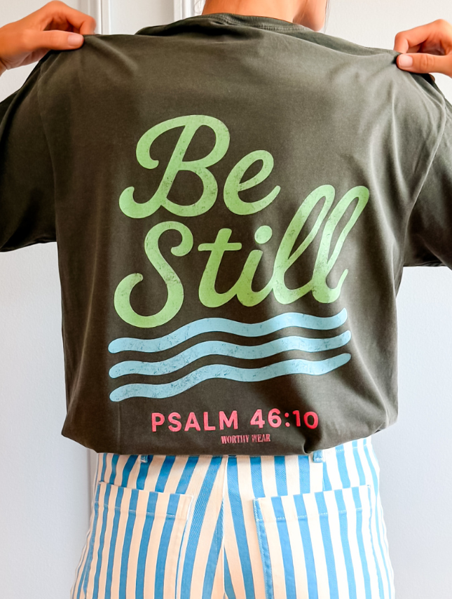 Green and pink "Be Still" graphic t-shirt with Psalm 46:10, worn by a person against a light wall.