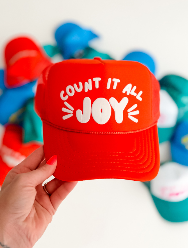 Red cap with white text saying "Count it all JOY," held in hand, surrounded by colorful caps in the background.