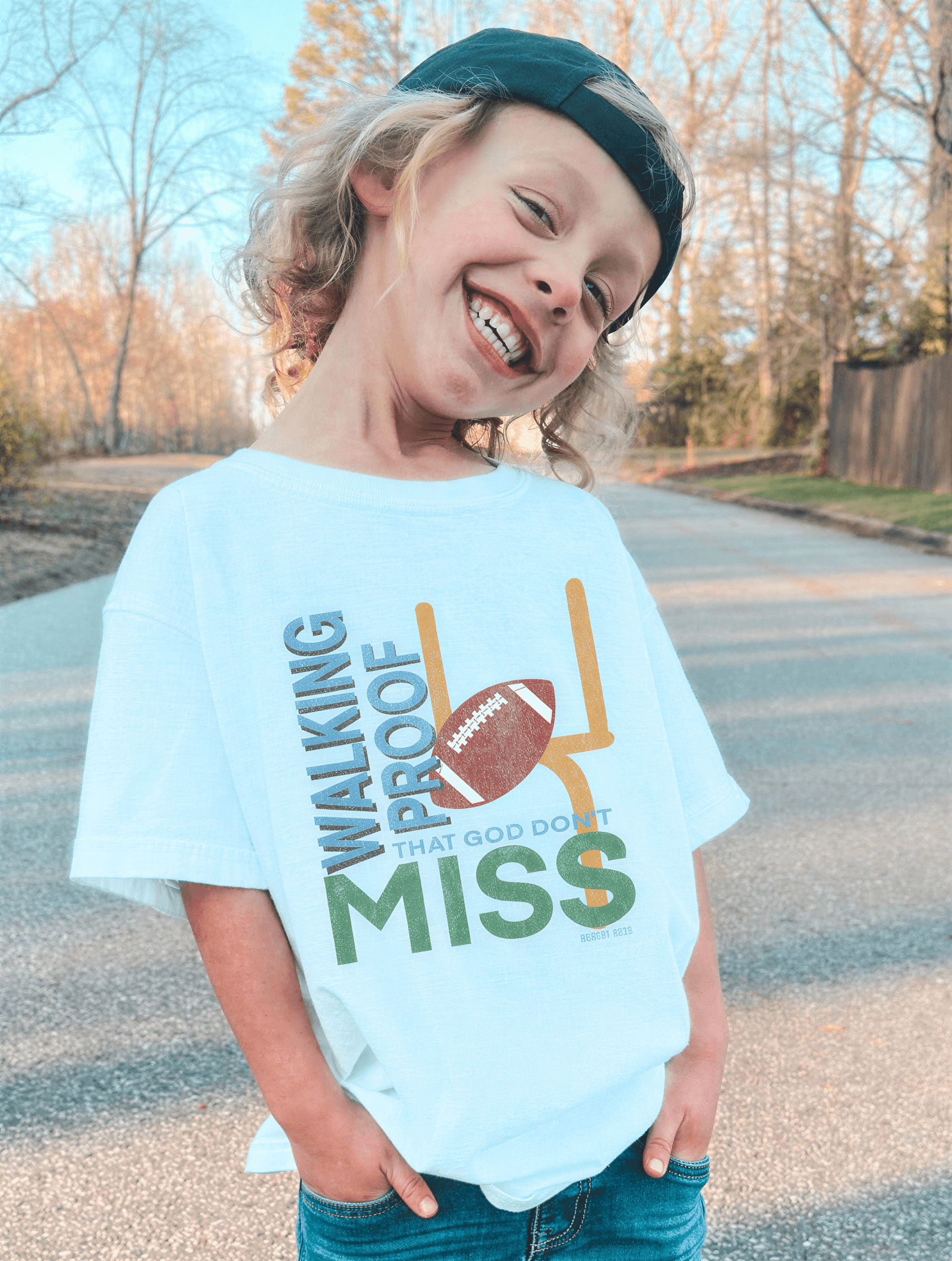 Child wearing a light blue t-shirt with a football graphic and text, standing on a road.