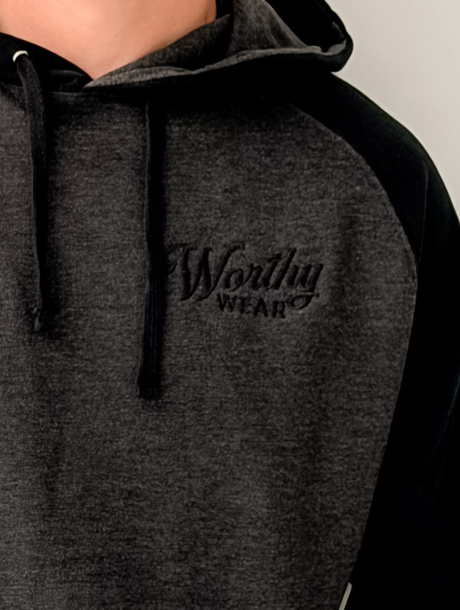 Two-tone hoodie in black and gray with embroidered "Worthy Wear" logo on the chest, featuring a drawstring hood.