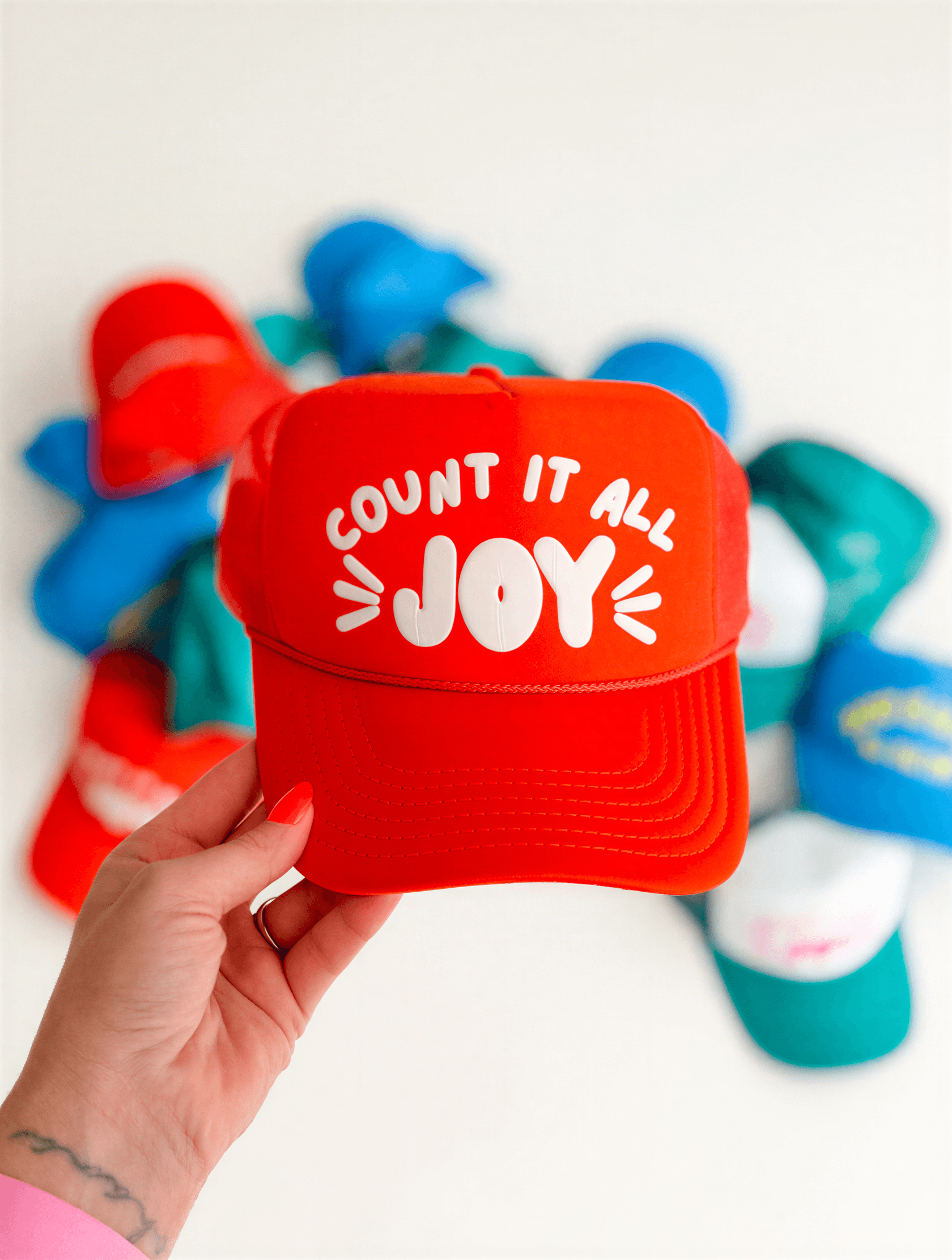 Count It All Joy foam trucker cap in orange, featuring white text on the front, held against a colorful background of caps.