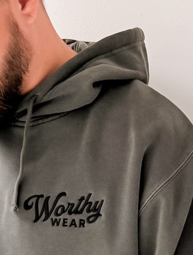 Worthy Wear washed hoodie in olive green with embroidered logo on the chest, worn by a person with a beard.
