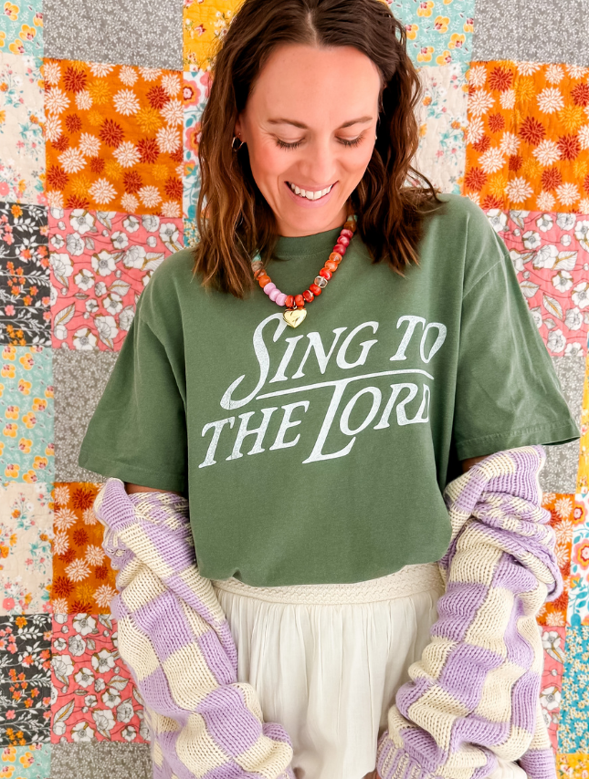 Green "Sing To The Lord" tee worn by a woman, paired with a pastel purple and white sweater, against a colorful quilt back...