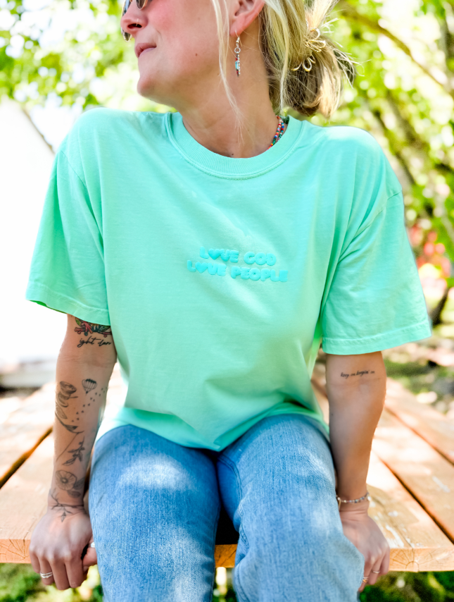 Mint green puff tee with "Love God Love People" text, worn by a person sitting on a wooden bench outdoors.