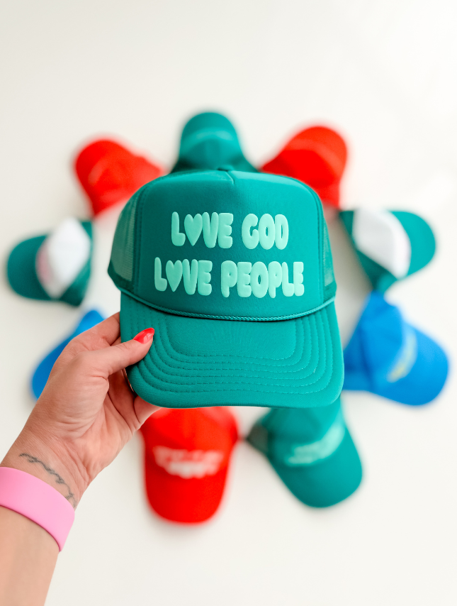 Turquoise foam trucker hat with "Love God Love People" text, held in hand, surrounded by colorful hats.