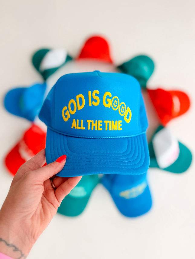 Blue foam trucker hat with "God Is Good All The Time" text, held in hand, surrounded by colorful hats in the background.