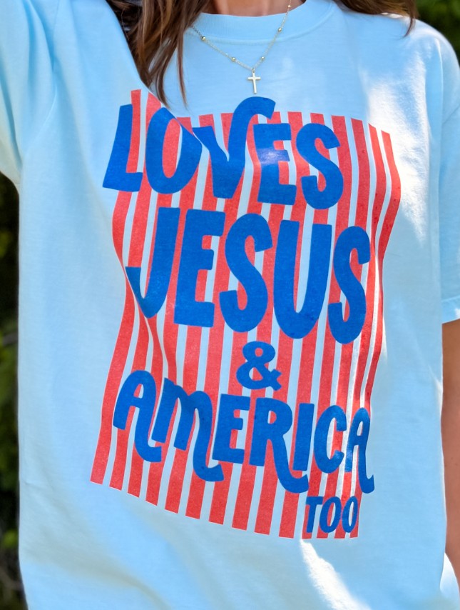 Loves Jesus & America tee in light blue with bold red and blue text on a striped background.