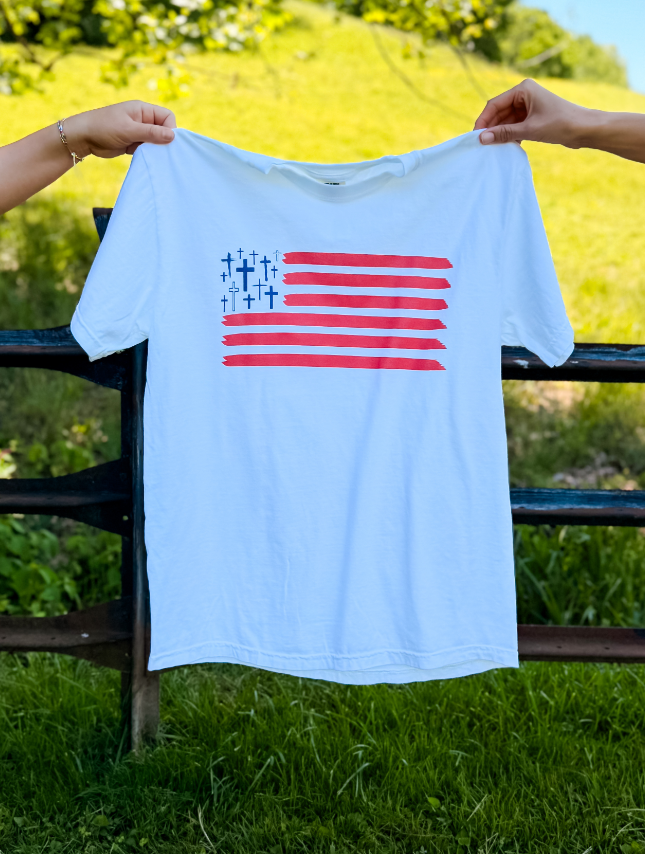 White Freedom In Faith tee featuring a red and blue striped design with crosses, held up against a green background.