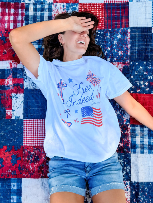 White Free Indeed tee with patriotic design, featuring a cross, fireworks, and an American flag, worn by a woman on a quilt.
