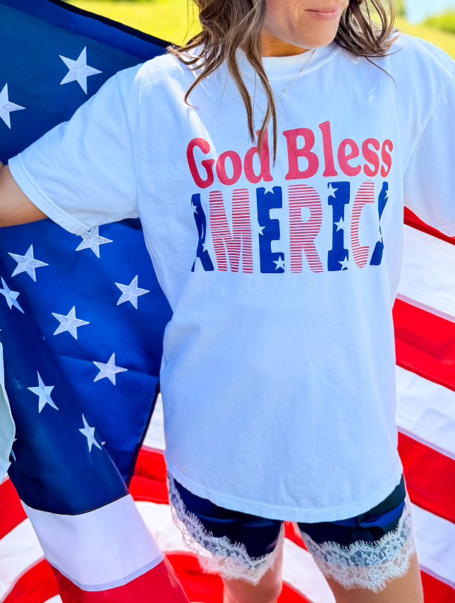 White t-shirt with "God Bless America" in red and blue letters, worn by a person holding an American flag.