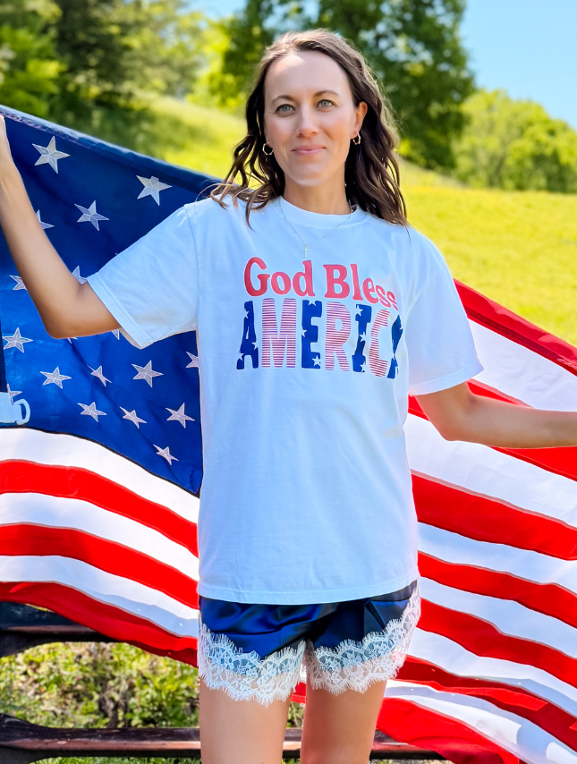 God Bless America tee in white, featuring bold red and blue lettering, worn by a woman holding an American flag.