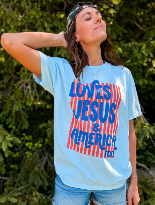 Light blue t-shirt featuring "Loves Jesus & America too" in red and blue text, worn by a woman outdoors.