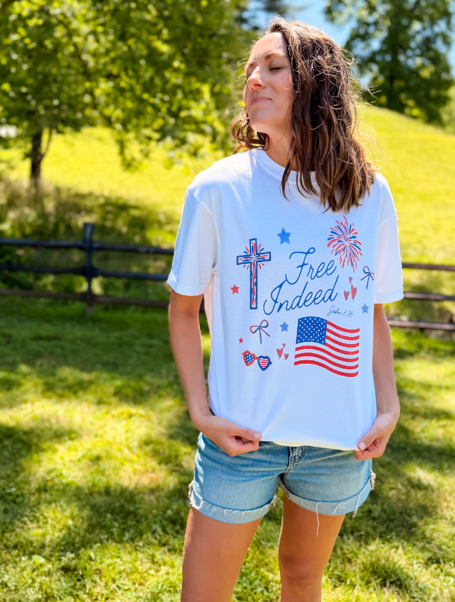 White short-sleeve tee featuring "Free Indeed" text, patriotic designs, and a model standing outdoors.