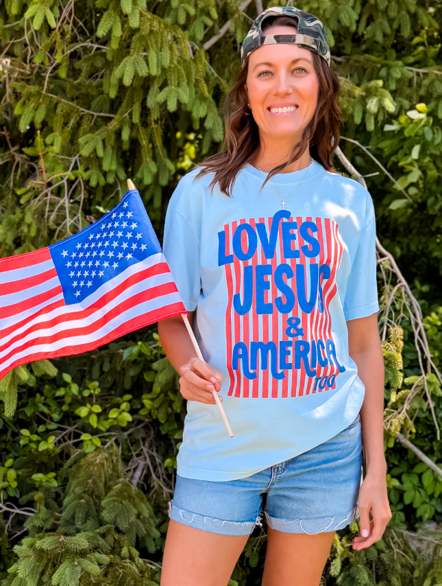 Loves Jesus & America tee in light blue, featuring bold red and blue text, worn by a woman holding an American flag.