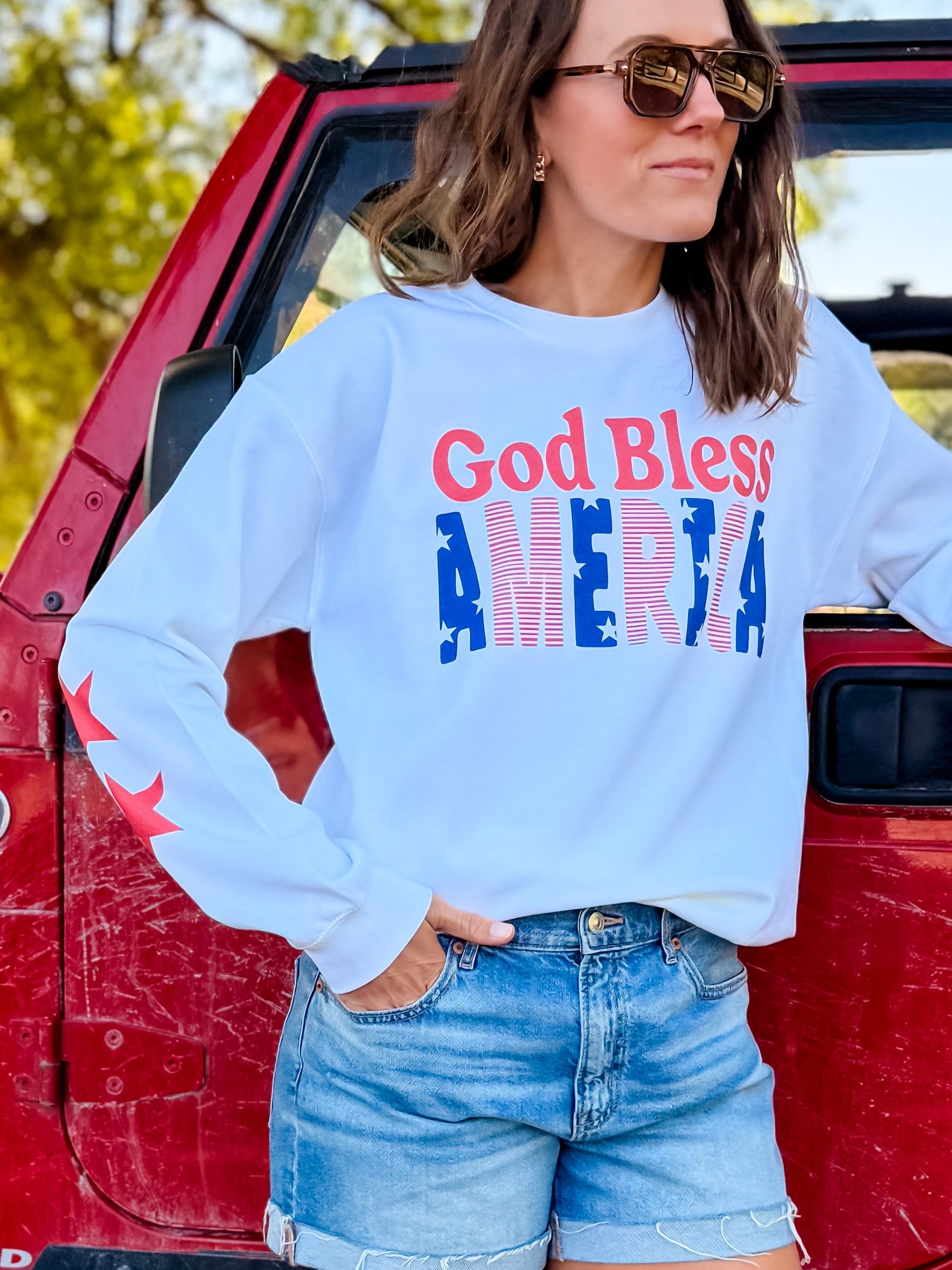 God Bless America sweatshirt in white with red and blue lettering, worn by a woman with sunglasses, standing by a red vehi...