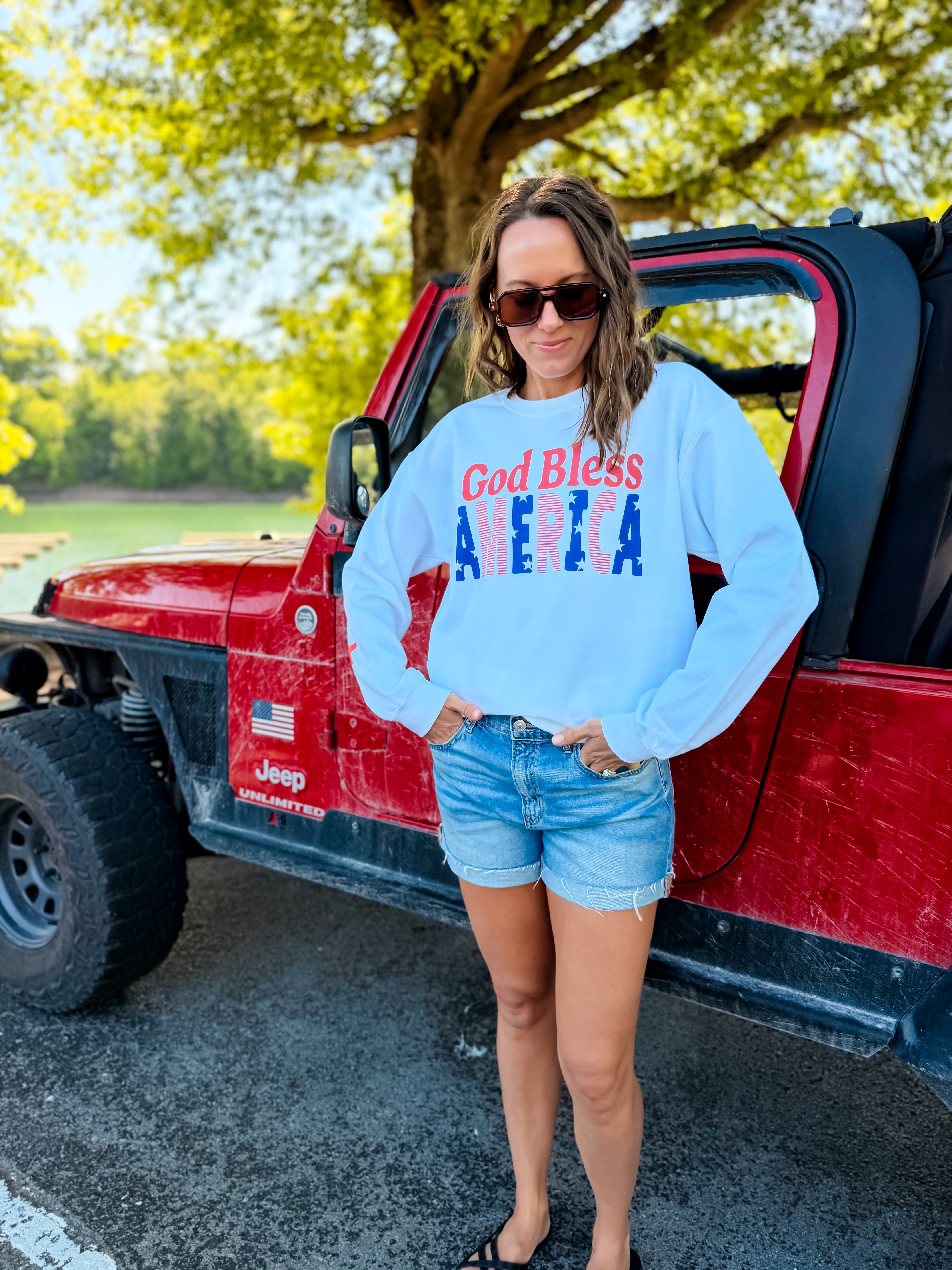 God Bless America sweatshirt in white, featuring red and blue lettering, worn with denim shorts near a red Jeep.